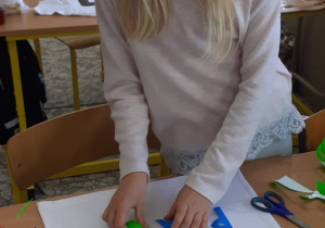 Dziewczynka pochyla się nad biurkiem i układa kompozycję z kolorowych liter na białej koszulce.